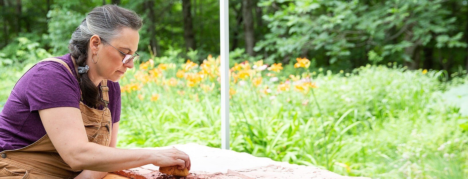 Val Gilman, artist and coach, dressed in studio overalls, working on art with garden and woods behind her.
